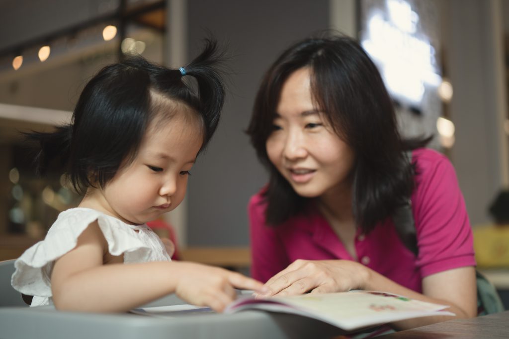 concept of Asian mother and 2 years old spending time together learning and doing activities, avoiding screen time by using paper book and sticker, while the daughter concentrate and enjoy learning.